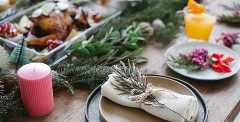 A festive holiday table setting with a plate and napkin adorned with a sprig of greenery, surrounded by decorations, a lit candle, a glass of orange beverage, and dishes of holiday food in the background.
